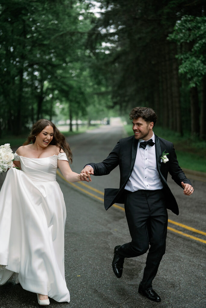 Bride and groom running together on wooded Michigan road during romantic editorial wedding session.