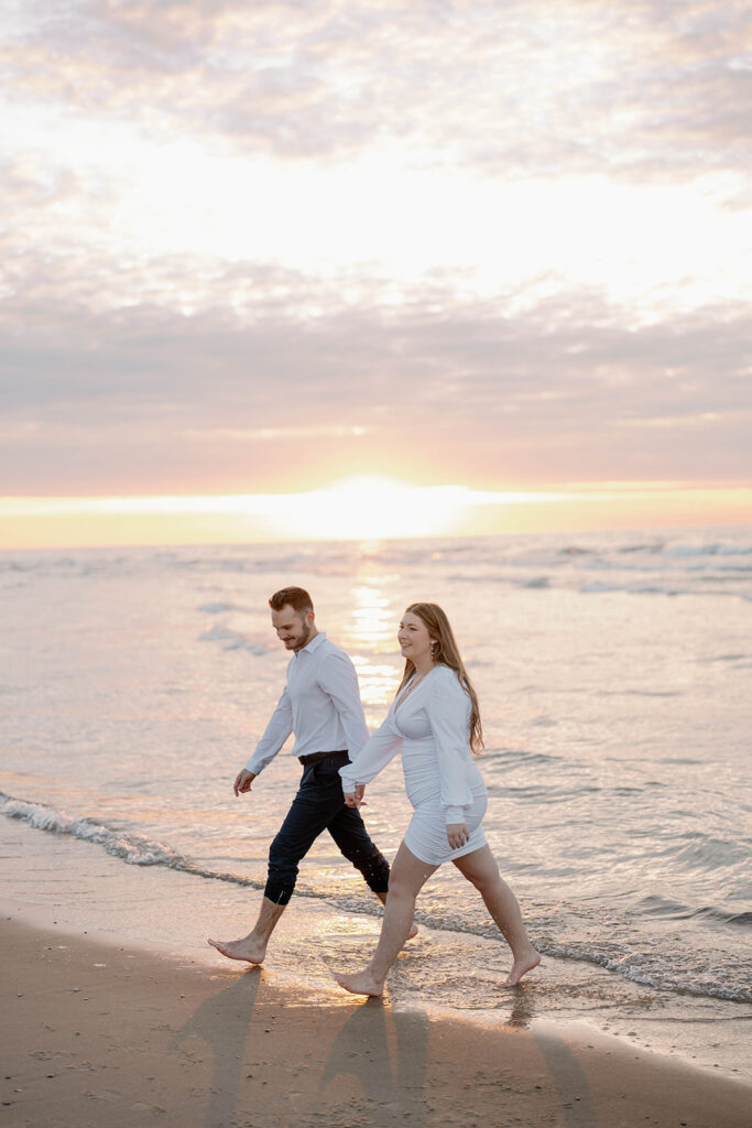 Engaged couple walking along shoreline of Lake Michigan, candid beach engagement photos at sunset.