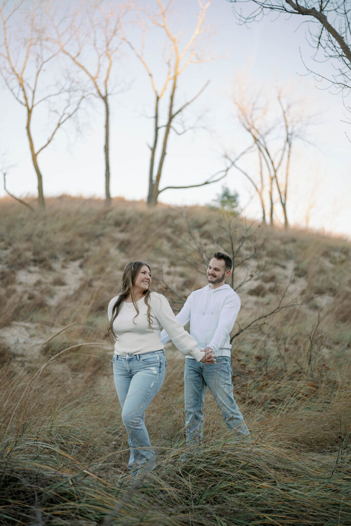 Couple walking through sandy grass near Lake Michigan dunes during engagement session, natural outdoor portraits.”