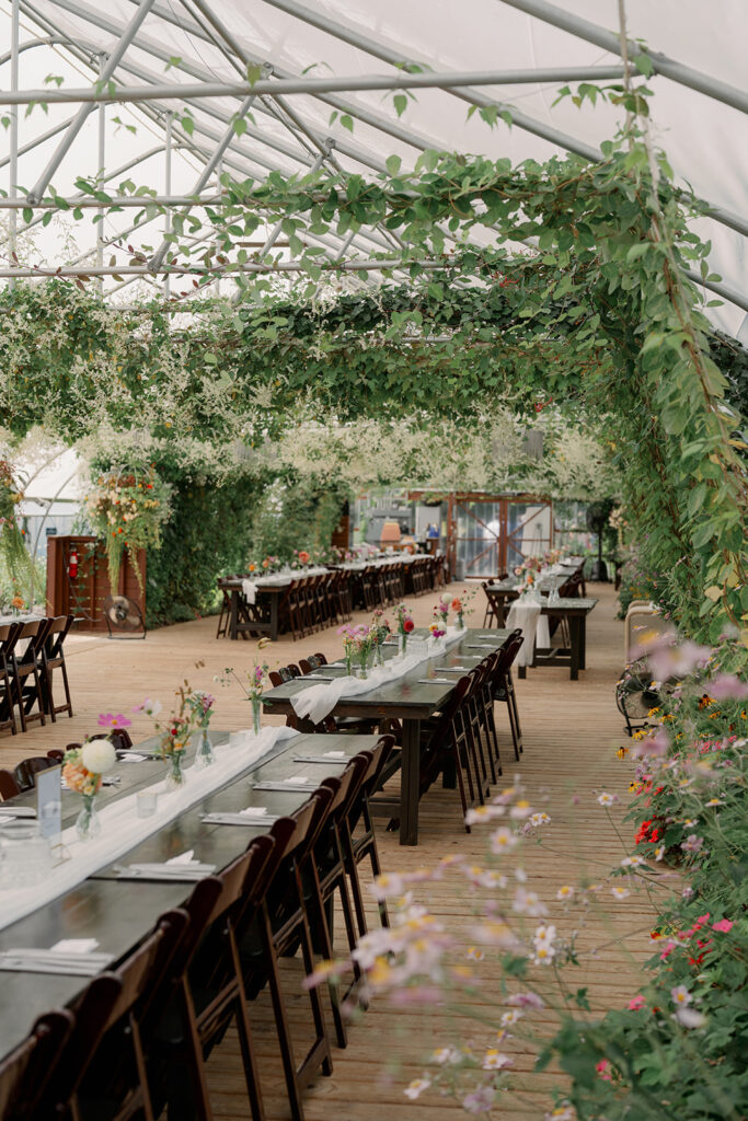 Reception inside the greenhouse at Of The Land with greenery hanging from the ceiling and long family style tables