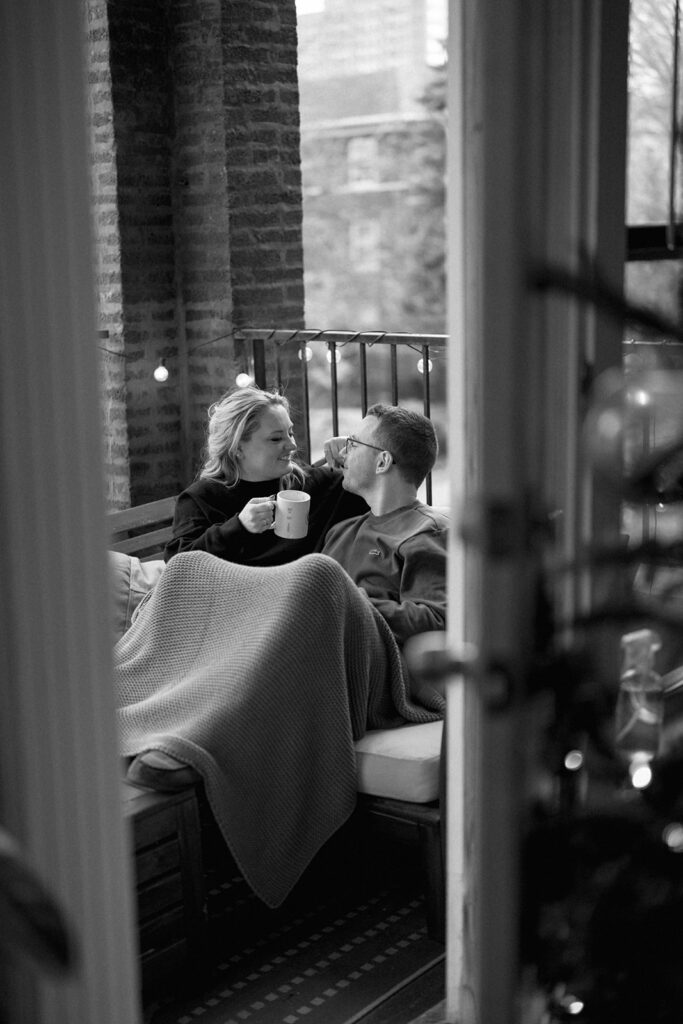 Black and white photo of engaged couple cuddling indoors during cozy lifestyle session.