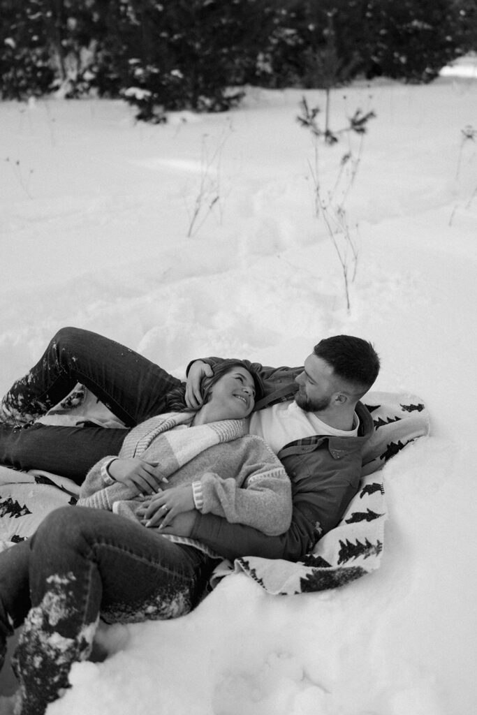 Black and white image of a couple lying in the snow laughing during a winter session in Kalamazoo MI