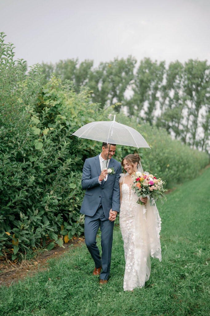 bride and groom walking through orchard under umbrella at Nugent Orchards wedding in Michigan